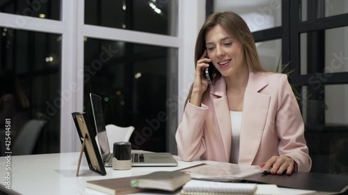 Confident businesswoman talking smartphone in office interior. Female professional looking at business documents during phone conversation. Business woman working with papers at remote workplace