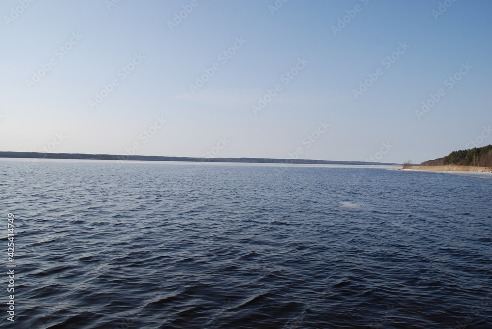 The undulating surface of a wide river under the spring sun. Blue water surface with small waves under a light blue sky. A path from the sun is reflected in the water. In the distance, a dark strip of