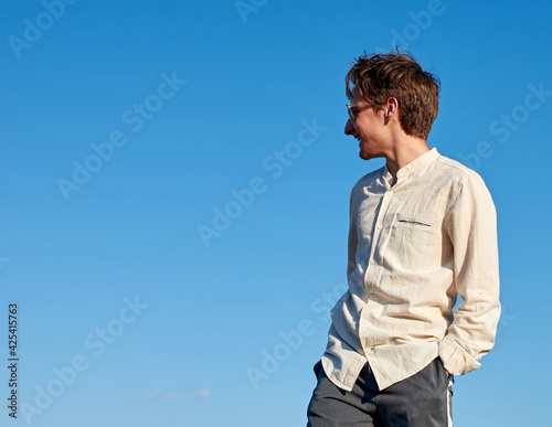 A Caucasian white man with his hands in his pocket looking to the side and smiling on sky background