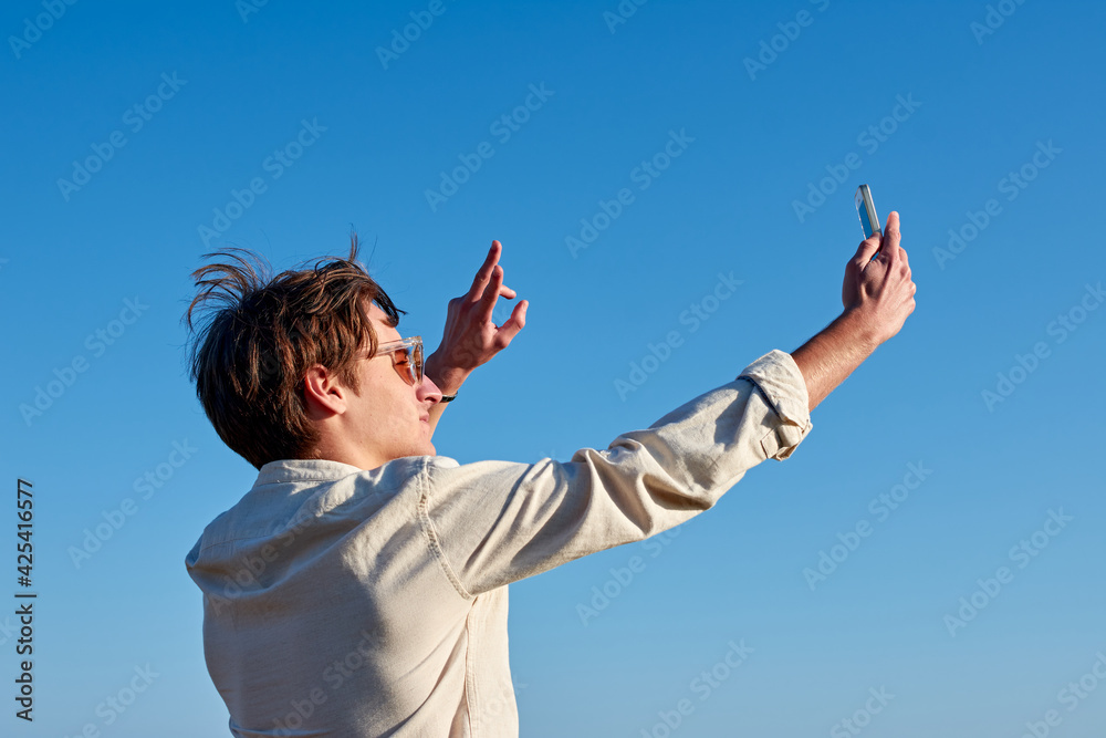 A Spanish white man with glasses and a beige shirt taking a selfie and making a victory sign on clear blue sky background A Spanish white man with glasses and a beige shirt taking a selfie and making a victory sign on clear blue sky background