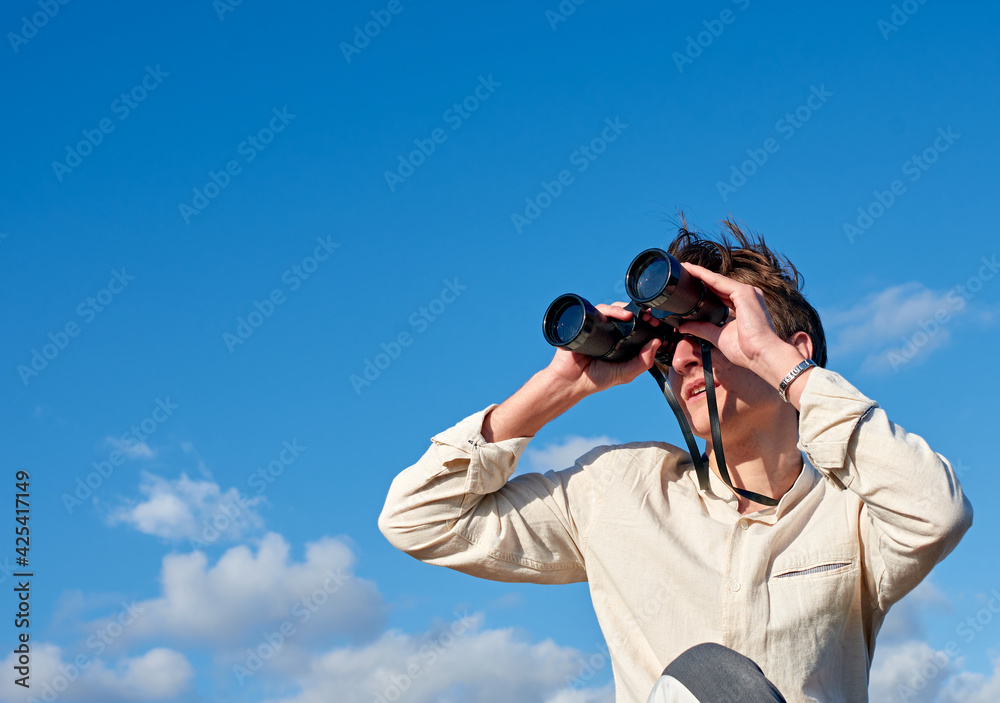 A Spanish white man in a beige shirt looking through binoculars on cloudy sky background A Spanish white man in a beige shirt looking through binoculars on cloudy sky background