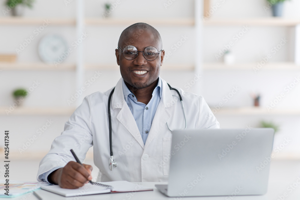 Friendly african american male doctor working with laptop in modern office, taking notes, planning his day