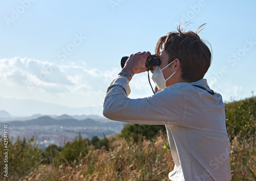 A shallow focus shot of a young man from Spain in a white shirt looking over a city through binoculars during the pandemic