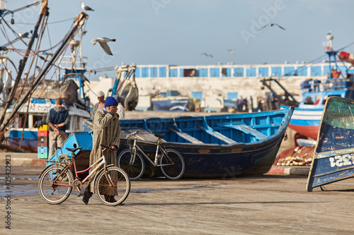 Man at fish market in Essaouira/Morocco pushing his bike and rubbing his eyes. Empty boat lies there. It is probably too early in the morning. 