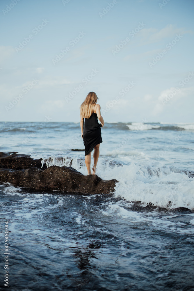 Photoshoot of a blonde girl in a dark dress on the Bali beach with black sand. photo by the cliff, photo by the big stones