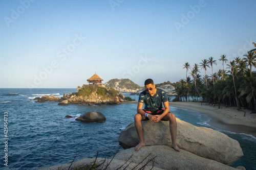 Hombre joven con gafas de vacaciones sentado en una roca junto al mar azul en un día soleado de verano  © hectorfabio