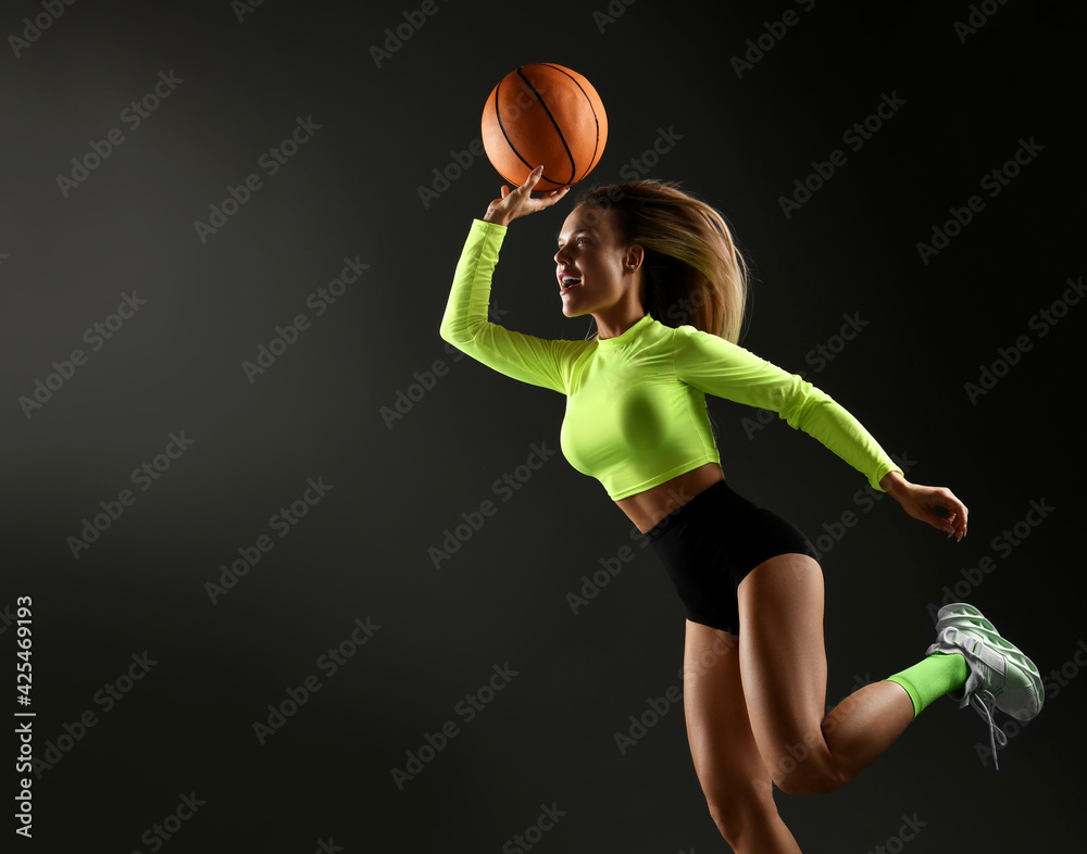 Young sexy blonde fitness woman in tight sportswear, top, shorts, high socks and sneakers jumps playing basketball ball over dark background with copy space. Female play basketball game