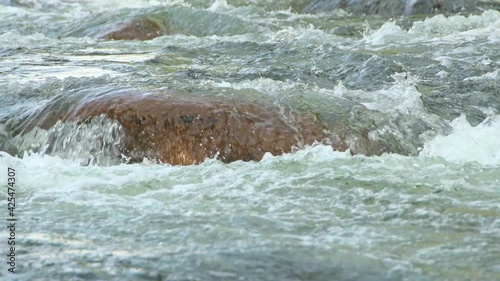 Turquoise water flows down granite boulder. River boils and forms waves. Drops splashes are reflected in sun