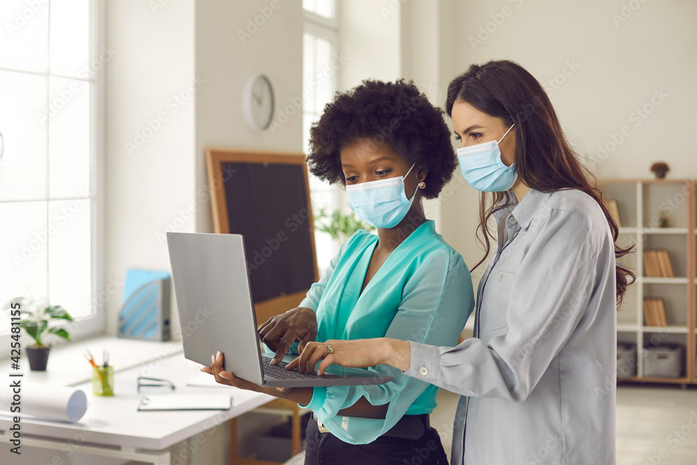 Fototapeta premium Two office coworkers in medical face masks using laptop together. Young business women discussing new project. Returning to work after Covid-19 lockdown. Safety measures during coronavirus pandemic