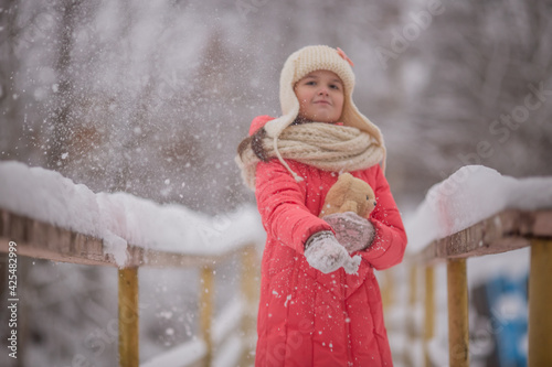 A 10-11-year-old girl walks through a snow-covered park. She's wearing a white knit hat and scarf. She's wearing a pink down jacket. She's having fun, throwing snow. She walks with a teddy bear.