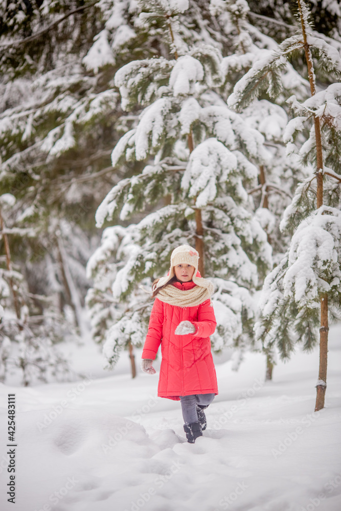 A 10-11-year-old girl walks through a snow-covered park. She's wearing a white knit hat and scarf. She's wearing a pink down jacket. She's having fun, throwing snow. She walks with a teddy bear.