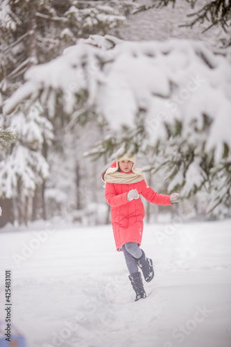 A 10-11-year-old girl walks through a snow-covered park. She's wearing a white knit hat and scarf. She's wearing a pink down jacket. She's having fun, throwing snow. She walks with a teddy bear.