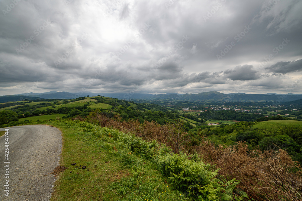 Landscape of the countryside of the french basque country, the sky full of clouds, in a cloudy day, below the green hills and meadows and to the left a curve of the road that disappears, in the backgr