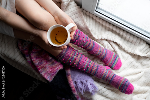 a young girl in lilac knitted socks-knee socks sitting on a white soft blanket on the windowsill with a cup of tea in her hands. Cozy handmade knitted things