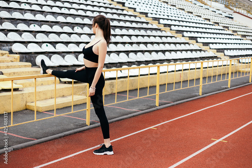 Young athletic girl stretches, prepares her body and muscles for a productive fitness workout treadmill race running track. Flexible female sporty model on the city stadium. Image with copy space. 