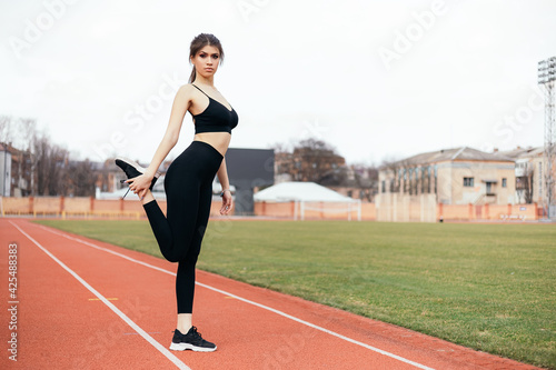 Young athletic girl stretches, prepares her body and muscles for a productive fitness workout treadmill race running track. Flexible female sporty model on the city stadium. Image with copy space. 