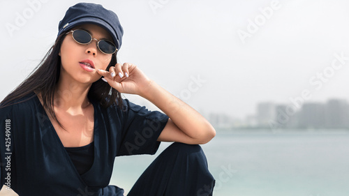 Outdoor lifestyle portrait of teen girl with amazing stylish outfit and sunglasses. Close-up portrait of natural beauty face with nude makeup. She bites her finger. Hand near the neck. Cityscape 