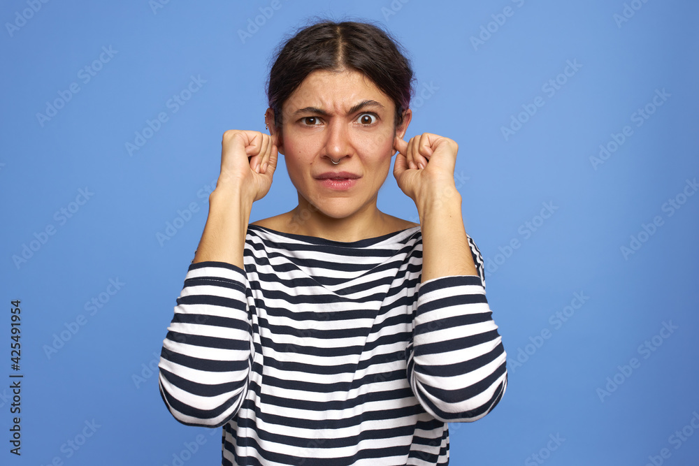 Human emotions, reactions, feelings and body language. Funny shocked young Caucasian woman in striped shirt plugging ears with fingers because of noise, looking at camera witt frustration, frowning Human emotions, reactions, feelings and body language. Funny shocked young Caucasian woman in striped shirt plugging ears with fingers because of noise, looking at camera witt frustration, frowning