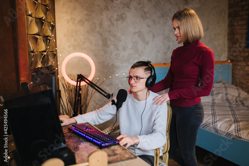 Young attractive wife help husband to work. Handsome man sits at the table and work on computer. Man using professional microphone and headphones
