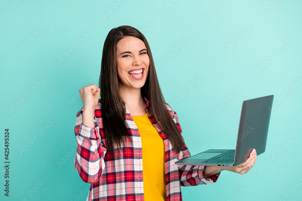 Photo of cheerful victorious young woman raise fist hold computer winner isolated on teal color background Photo of cheerful victorious young woman raise fist hold computer winner isolated on teal color background