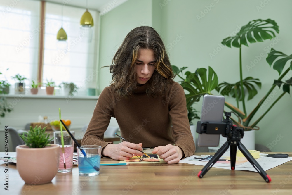 Teenage guy drawing with watercolors, watching video class on smartphone