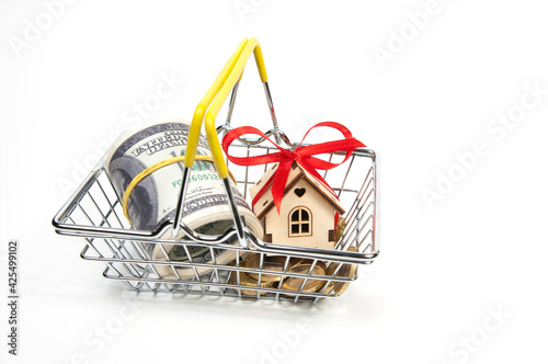 in the shopping basket at the supermarket, a rolled-up bundle of hundred-dollar bills, coins and a house with a red bow on a white background is isolated, the concept of buying a real estate, a house.