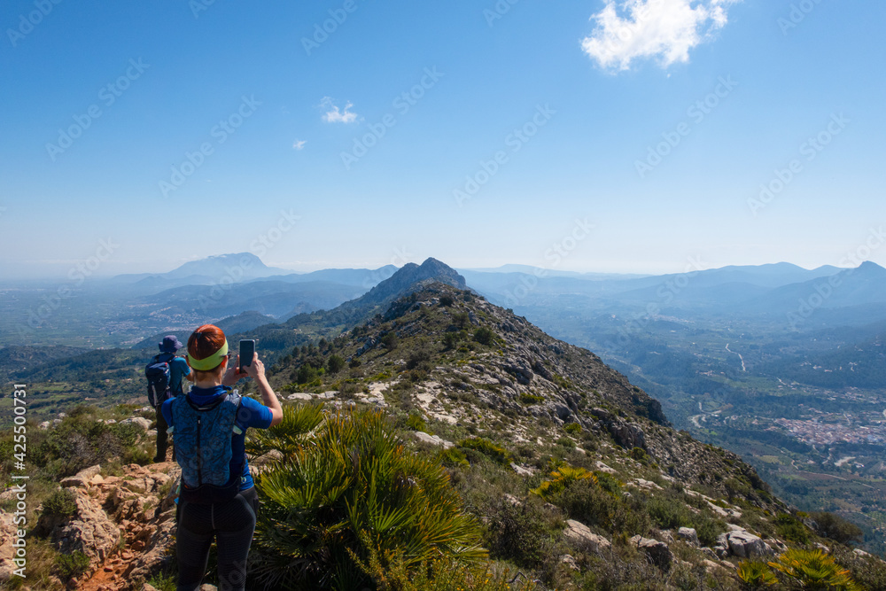 Hiker on a mountain, taking photos with the smartphone, with a clear sunny sky.