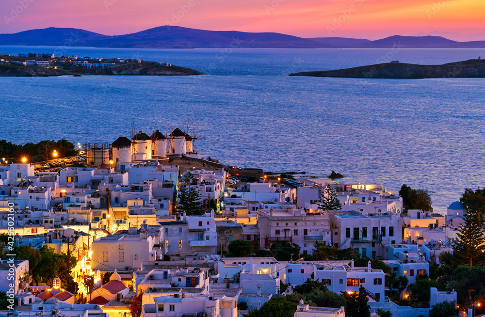 Beautiful sunset view of famous traditional white windmills, Mykonos, Cyclades, Greece. Whitewashed houses, colorful sky, summer evening, town lights