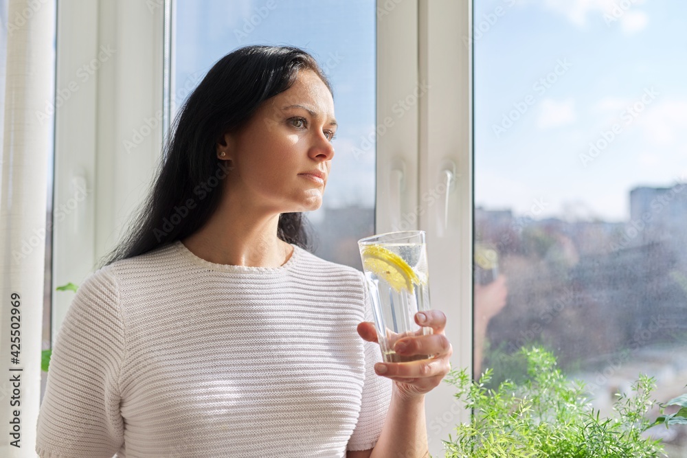 Beautiful woman with glass of water with lemon at home near window Beautiful woman with glass of water with lemon at home near window
