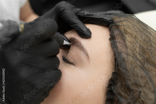 Concept of microblading eyebrows. The cosmetologist performs the procedure of permanent makeup of the eyebrows in close-up.