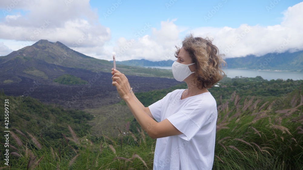 Side view of young Caucasian woman in a face mask taking pictures of mountains with her smartphone. Solo travelling during covid-19 pandemic