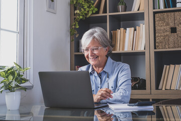Happy old caucasian businesswoman smiling working online watching webinar podcast on laptop and learning education course conference calling make notes sit at work desk, elearning concept
