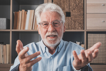 Head shot portrait confident old businessman coach wearing glasses looking at camera and talking, mentor speaker holding online lesson, explaining, sitting at wooden work desk in modern cabinet
