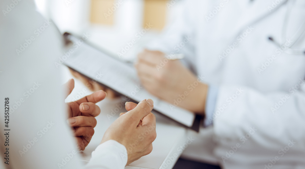 Unknown male doctor and patient woman discussing current health examination while sitting in clinic and using clipboard. Perfect medical service in hospital. Medicine and healthcare concept Unknown male doctor and patient woman discussing current health examination while sitting in clinic and using clipboard. Perfect medical service in hospital. Medicine and healthcare concept
