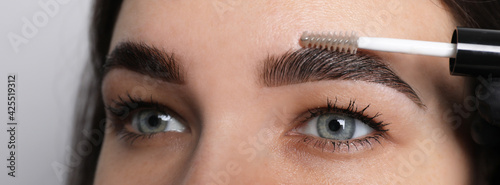 Beautician fixing woman's eyebrows with gel after tinting on light grey background, closeup photo