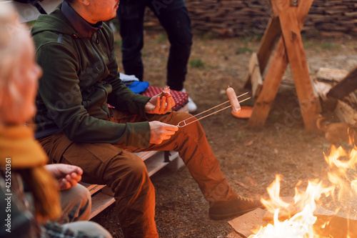 Grilling sausages over a campfire, campers roasting sausages on toasting forks. Fire place, friends, tourists are sitting near the flame.