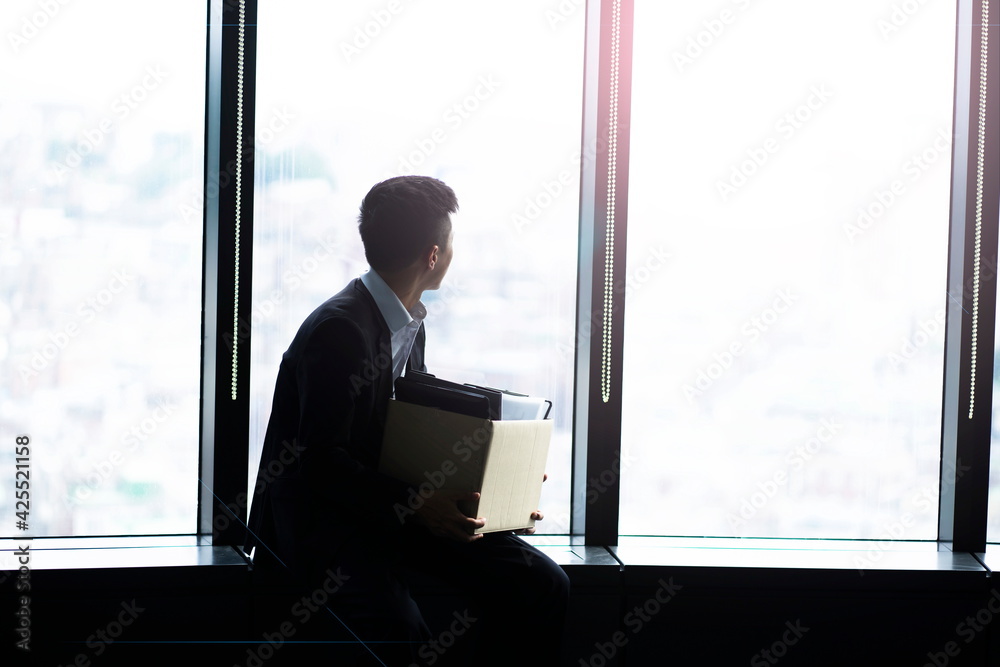 Fired businessman sitting with box of belongings by window
