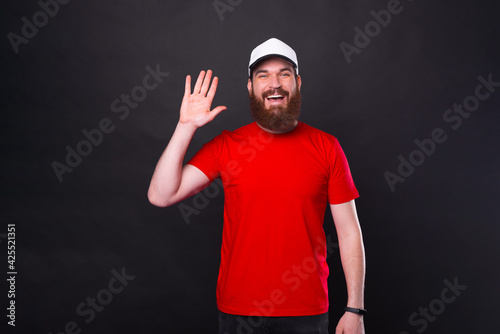 Portrait of smiling cheerful young bearded man in red t-shirt saying Hello over black background