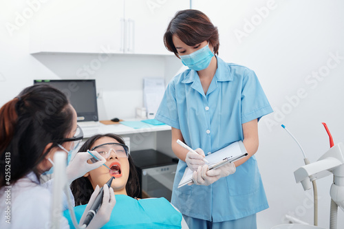 Dentist examining teeth of female patient when nurse taking notes in document