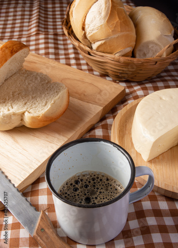 Breakfast table in Brazil with breads, cheese, cup of coffee and accessories on a brown and beige checkered tablecloth, dark background, selective focus.