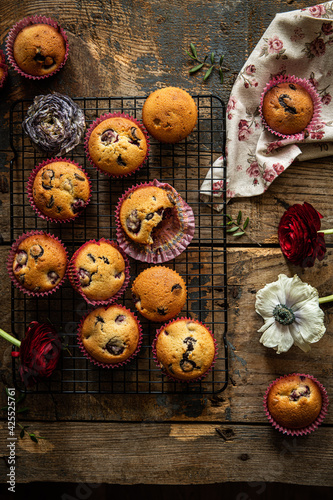 Group of homemade cherry and chocolate muffins on cooling rack and fresh flowers and leaves on rustic wooden table.
