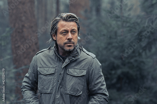 Blonde man in green coat walks in a pine forest.