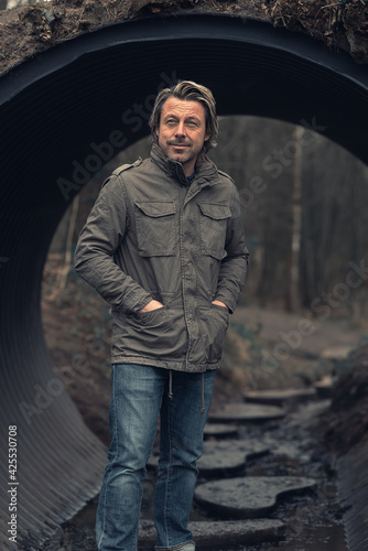 Blond man in green jacket and jeans stands in a tube tunnel in nature reserve.