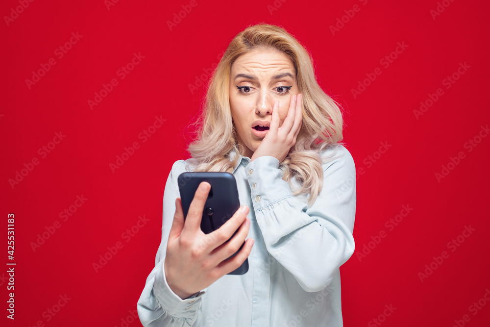 Astonished girl staring to phone, isolated on red background. Trendy woman. Unbelievable message. Studio shot Astonished girl staring to phone, isolated on red background. Trendy woman. Unbelievable message. Studio shot