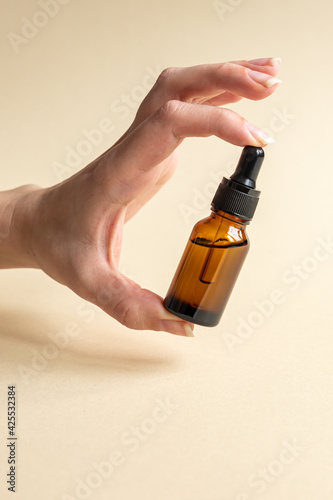 female hand with Dropper of essential oil, aromatherapy essence, beuty serum or medicinal liquid on beige background. Unbranded bottles for your design.