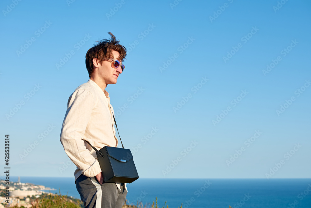 A Caucasian man from Spain standing on a grassy hill next to the ocean with a black purse over his shoulder A Caucasian man from Spain standing on a grassy hill next to the ocean with a black purse over his shoulder