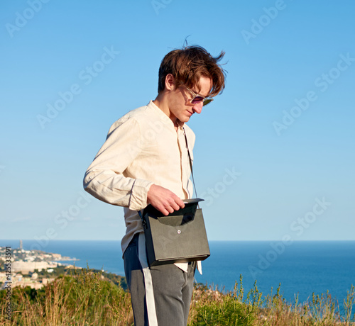 A Caucasian man from Spain standing on a grassy hill next to the ocean and closing his black handbag © Érik Glez.