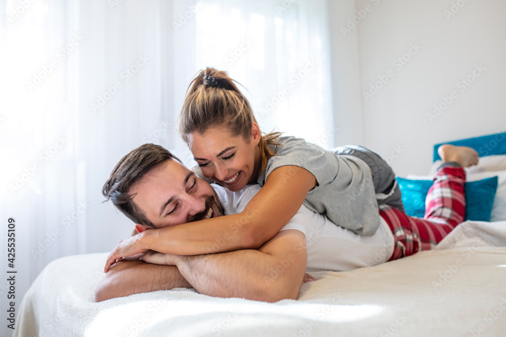 Beautiful young couple in bedroom is lying on bed. Enjoying spending time together. Smiling tender couple hugging in bed in the morning. Couple in love wearing pajamas cuddling while lying in bed
