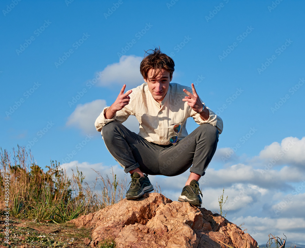 A Caucasian man from Spain making a peace sign with both hands while squatting on a rock surrounded by grass A Caucasian man from Spain making a peace sign with both hands while squatting on a rock surrounded by grass