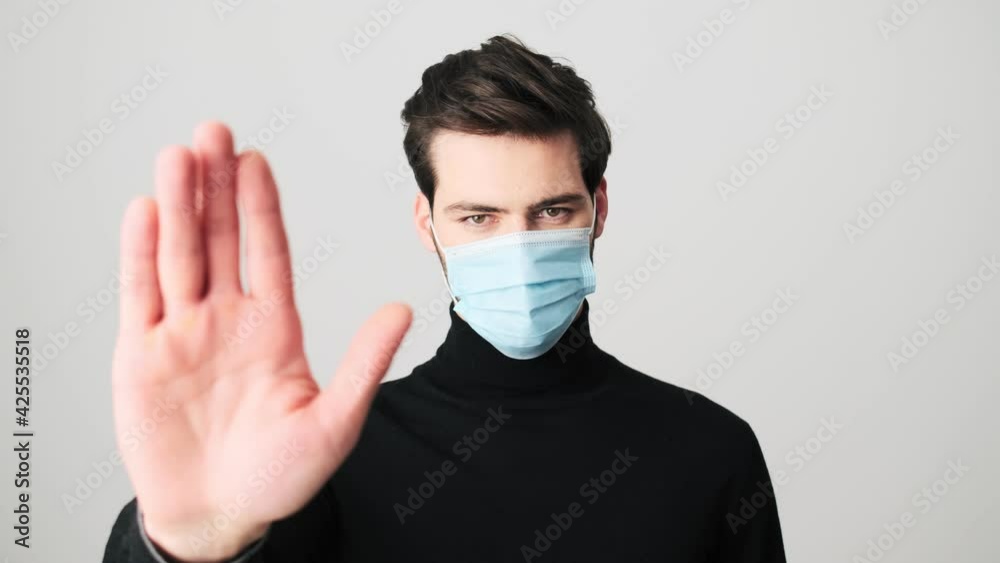 A serious man with a protective medical mask is doing stop gesture standing over white background